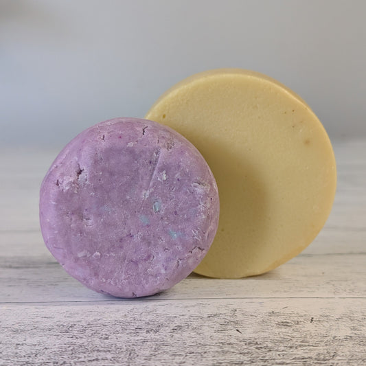 One shampoo bar and one conditioner bar, one purple and one yellow, on a wooden surface with a blurred background.
