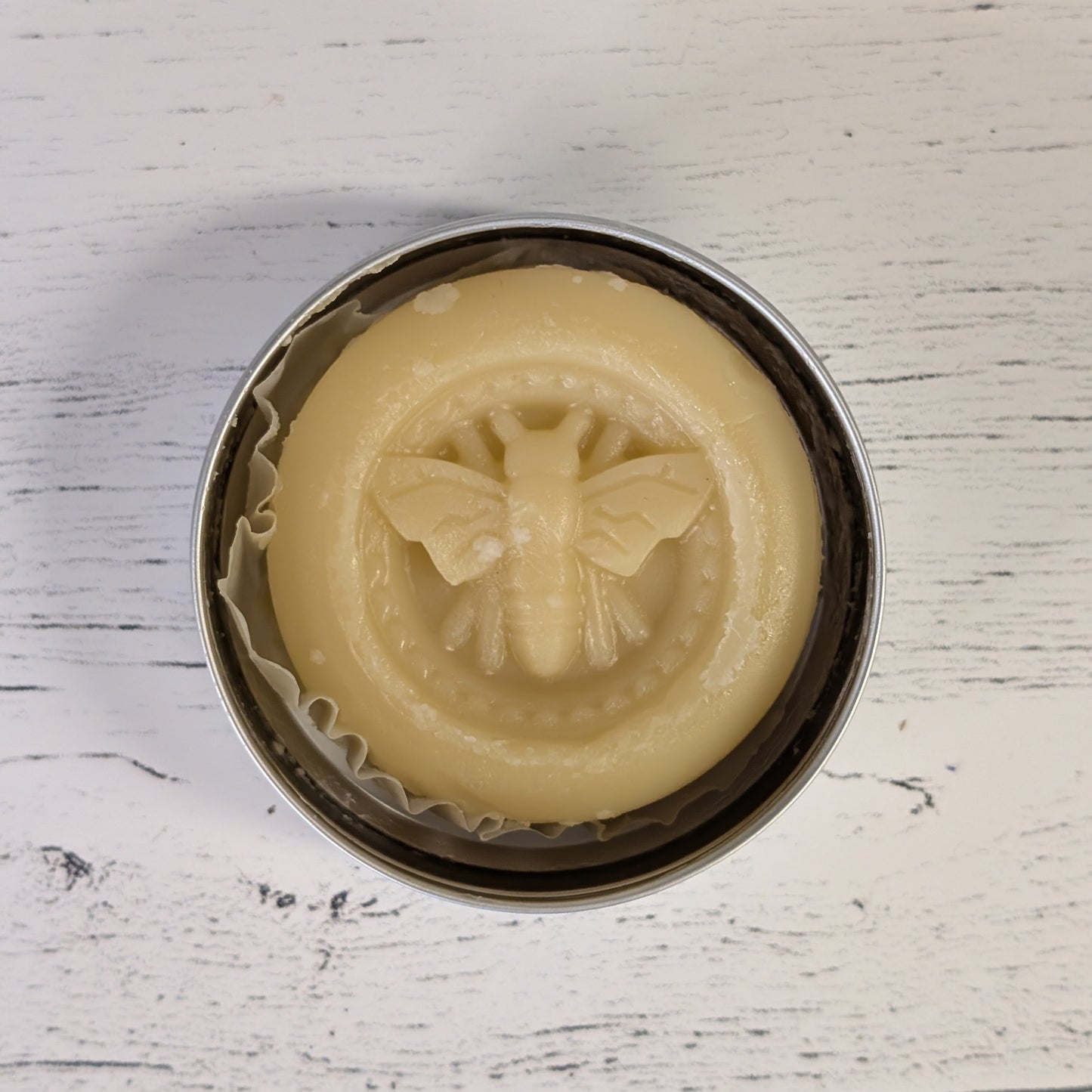 Beige soap with bee design in a metal tin on a white wooden surface